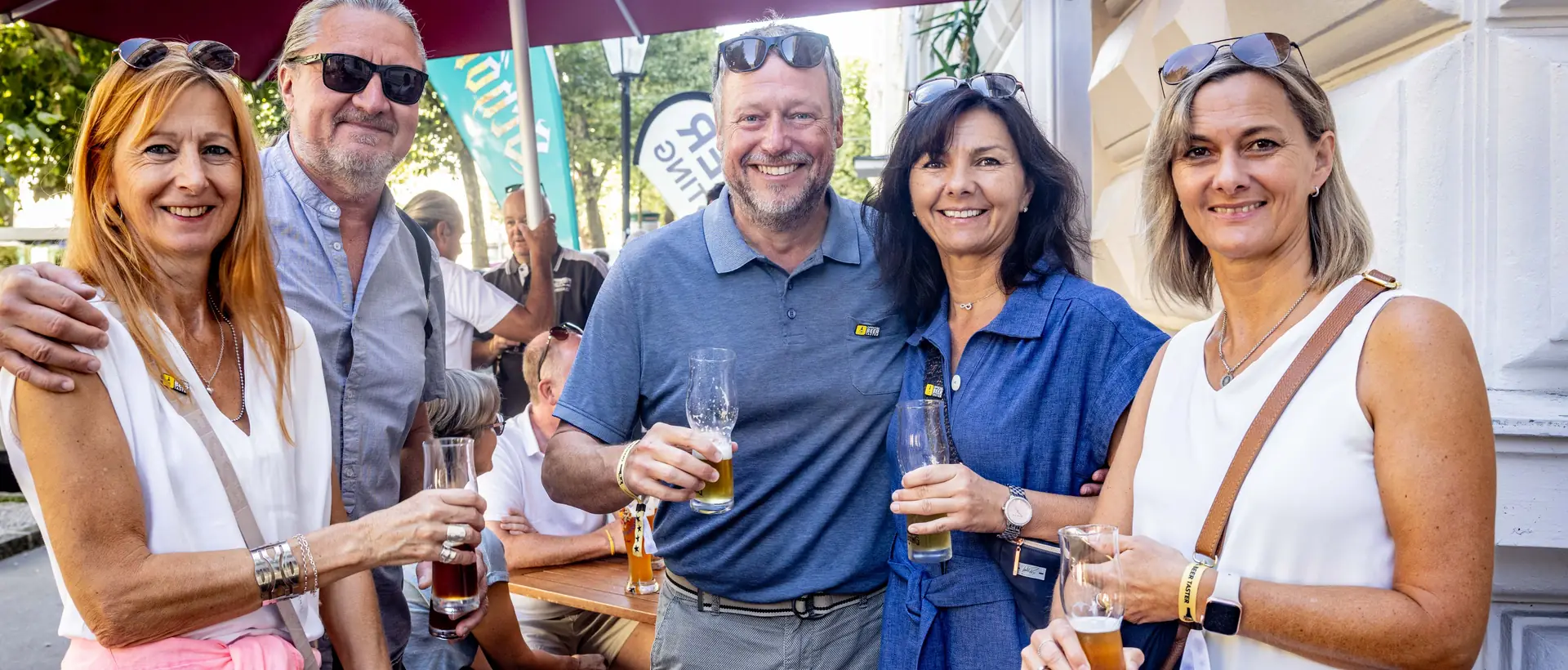 Fünf Gäste stehen lachend mit Biergläsern vor einem Lokal in der Sonne