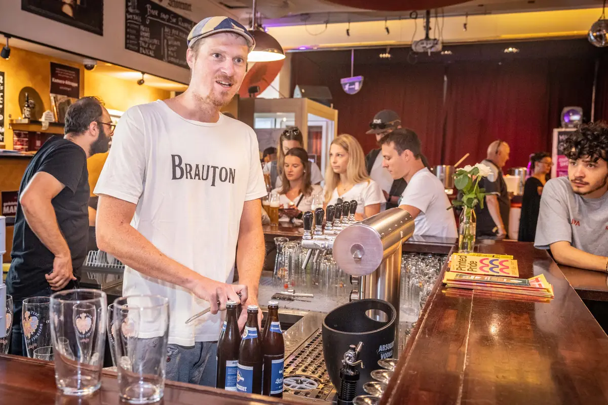 Brauer im BRAUTON-Shirt öffnet Bierflaschen hinter der Bar, im Hintergrund Gäste