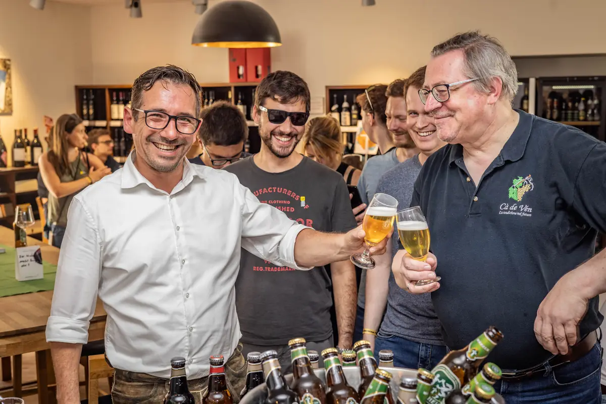Gruppe stößt in einem Bierladen mit Gläsern an, im Vordergrund eine Auswahl an Flaschen